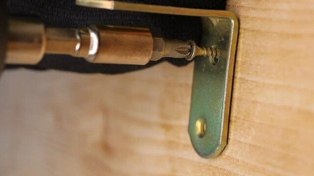 A brass corner is screwed with brass self-tapping screws to a wooden board using an electric screwdriver, close up.