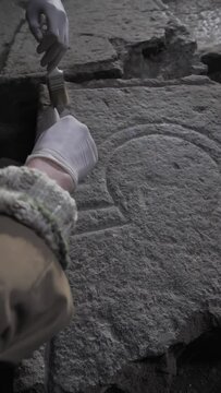 Vertical: Hands in gloves, two archaeologists using brushes sweep away dust from the diaracal sign of Libra carved on stone in an ancient temple. Astrological ancient symbols, alchemy designations