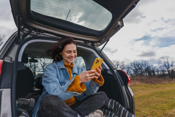 Young happy woman sitting in an open car trunk taking pictures with a camera. Traveling by car, communication in travel concept - 744518733