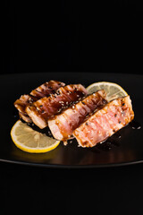 baked tuna sliced steak sprinkled with unagi sauce with lemon on a dark background in a black plate