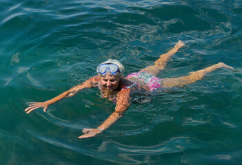 Aged woman is swimming with goggles in green sea water.