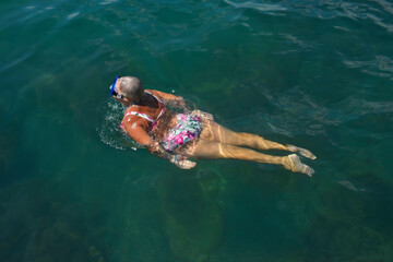 Aged woman is swimming with goggles in green sea water.