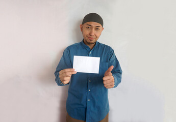 Adult Asian muslim man showing thumbs up while holding white blank envelope with happy expression