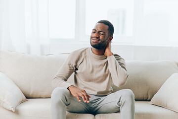 Stressed African American man suffering from a severe headache while sitting alone on a couch in his apartment His tired and sick expression reflects the physical and mental pain caused by the - Powered by Adobe