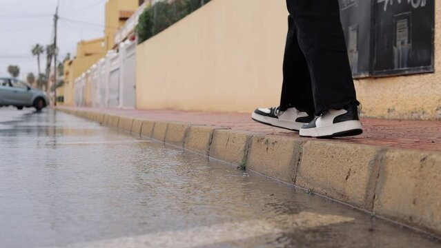 Female feet in black and white sneakers jump over a large puddle on the sidewalk on the street during the day, long close-up. Splashes fall on the affected parties. side plan.