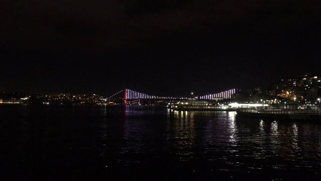 Walking along the Bosporus at night towards the 15 July Martyrs Bridge (15 Temmuz Şehitler K&ouml;pr&uuml;s&uuml;) at night. The bridge is lit up in the distance. A ferry maneuvers into the terminal. 