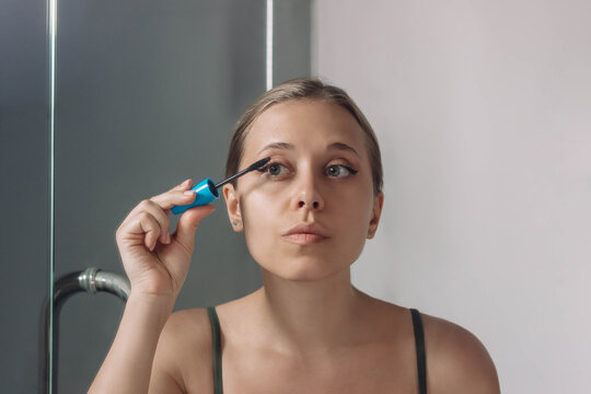 Young blonde woman using a brush applying black mascara to the eyelashes looking in the bathroom mirror. The result of lengthening and increasing the volume of natural lashes. Applying makeup