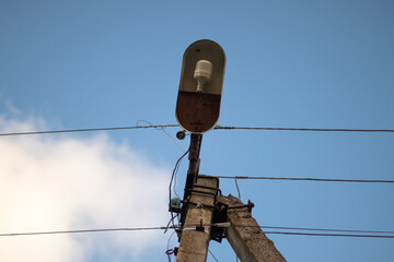 Old concrete lighting pole with a single lamp and wires against the sky with fluffy white cloud....