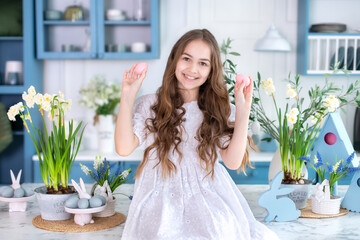 CloseUp portrait of happy little girl with easter eggs in hands and bouquet flowers in kitchen. Girl and flowers. Child is preparing for Easter.  Little Girl hunts and picks up easter eggs from table.