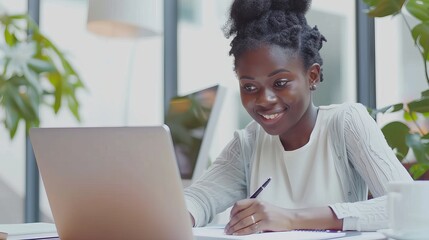 Business and education concept, a smiling young black woman sits at a desk working on her laptop and writing a letter in her paper notebook, free copy space. Happy millennial female studying on a