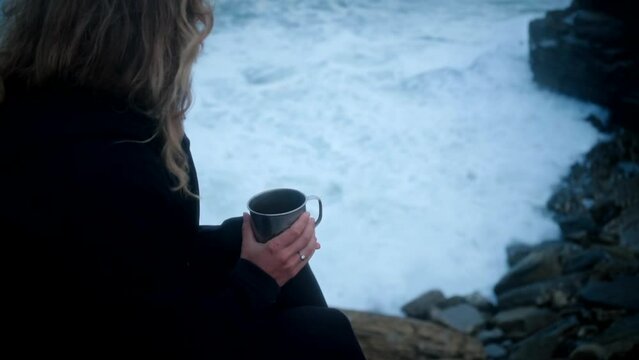 Woman sitting alone on the edge of a cliff, drinking tea or coffee and watching powerful ocean waves crashing on the shore. Moody atmosphere. Mental health concept. Space for text. 