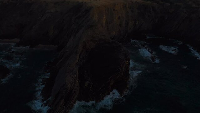 Camera flies over rocky cliffs in dark sunset dusk before night time. Ocean waves crashing on the shore mountain into the white foam. Beauty and power of nature. 