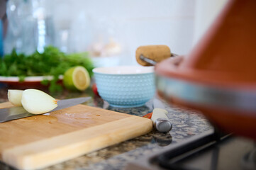 Cropped view of halves of an onion on a wooden board on the kitchen counter and a clay pot tagine steaming on the stove