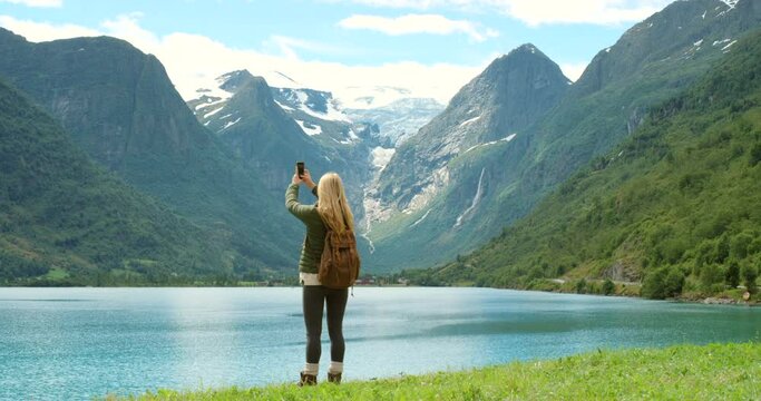 Woman, lake and cellphone for pictures, mountain and norway with backpack, view and sightseeing. Technology, exploring and nature for vacation, adventure and travelling in outdoor for tourism