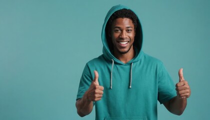A cheerful man in a teal hoodie gives two thumbs up, showcasing a relaxed and happy demeanor against a teal background. - obrazy, fototapety, plakaty