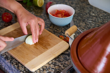 Cropped view of the hands of a chef cutting onion in two halves on a wooden board, using a kitchen knife.