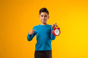 Child boy holding alarm clock and pointing with finger at it, over yellow background. Time...