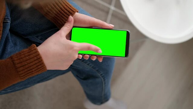 Young woman sitting at home holding smartphone green mock-up screen in hand. Female person use chroma key mobile phone. Vertical mode. Touching, swiping display, tapping, surfing internet social media