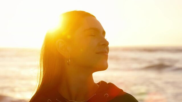 Head and shoulders shot of casually dressed young woman sitting on sand watching beautiful sunrise morning over beach and sea in South Africa with flaring sun  - shot in slow motion
