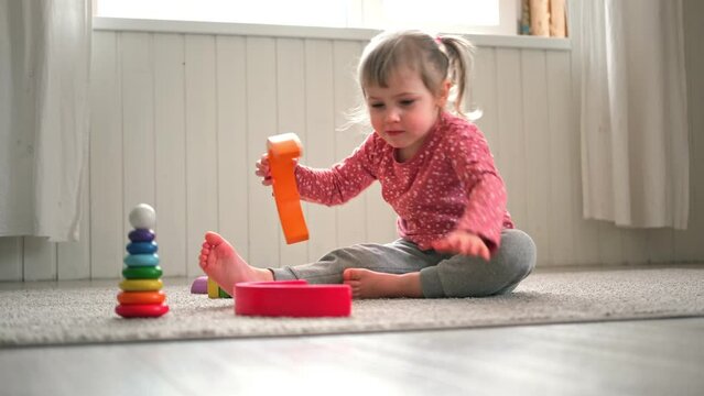 Kid girl play toys at home, kindergarten or nursery. Baby child playing playthings cubes and wood puzzle, pyramid sitting on the floor. Happy preschool education indoor. 3 years old little toddler.	

