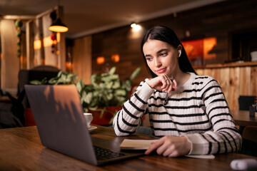 Woman in earphones using laptop while sitting at cafe. Young female student sitting in a coffee shop and working on laptop, looking conference.