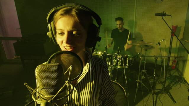 Singer, accompanied by live music, records song or soundtrack for film. Drummer plays drum kit, female professional vocalist sings in sound studio. Pop star performes hit for album