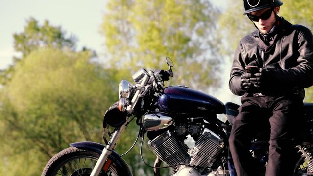 male motorcyclist in a leather jacket and helmet with a smartphone and chopper in the summer. Mobile application for motorcyclists.
