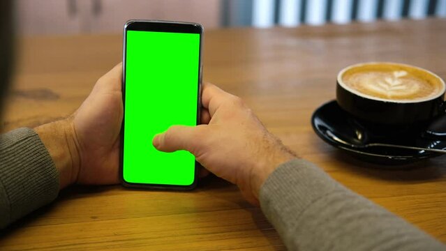 Young man sitting at cafe holding smartphone green mock-up screen in hand. Male person using chroma key mobile phone. Vertical mode. Touching, swiping display, tapping, surfing internet social media	
