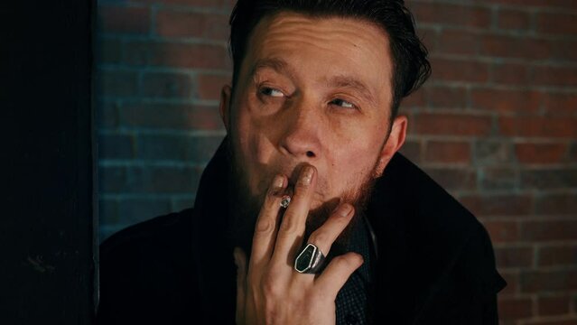 Bearded man is standing around corner of house, smoking, watching someone in late evening. Bandit takes puff while waiting for victim at night. Crime, danger, killer, thief, gangster, burglar