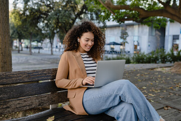pretty curly woman sitting in city street in stylish jacket, working remote job on laptop