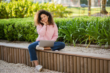 pretty curly young woman sitting in park working on laptop remote job