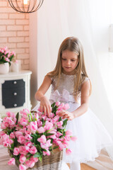 Little girl is standing in the bedroom. Large bouquet of tulip flowers. Princess in a white dress with a bouquet of pink flowers in the room.