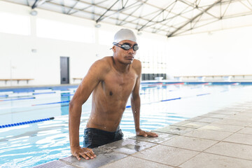 Young biracial male athlete swimmer takes a break at the poolside, with copy space