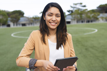 Young biracial teacher smiles holding a tablet on a sports field