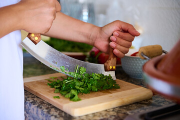 Close-up chef's hands using mezzaluna knife, chopping fresh green herb of parsley on a bamboo cutting board