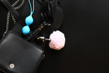 Sex toys. A small black bag with vaginal balls, a metal anal plug, a vibrator and a leather whip for intimate games. Women's objects for sexual entertainment on a black background - obrazy, fototapety, plakaty
