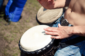 Man playing a djembe drum without a shirt outdoors on grass