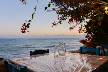 Bottle and transparent glass jug on table in typical Greek tavern at sunset with the Aegean Sea in the background