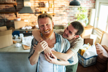 Fototapeta na wymiar Gay male couple hugging and laughing in new home with moving boxes