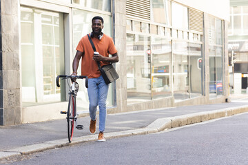 A young African American man walks with his bike outdoors in the city with copy space