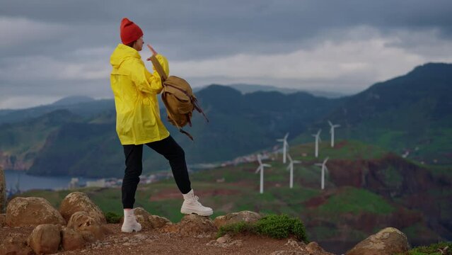 Attractive traveling woman in a yellow jacket and backpack looking at windmills in field 