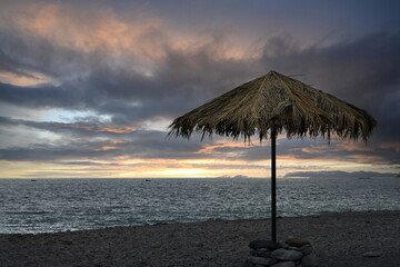 A lone beach umbrella with a covering of withered palm leaves on a shingle shore of the Atlantic...