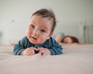 A three-month-old boy lies on his stomach on the bed and his mother sleeps behind him. Postpartum...
