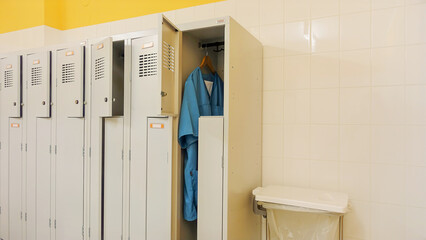 Open metal lockers in the locker room. A nurse's clothes, turquoise smocks and trousers, are hanging on a hanger. Concept of acute shortage of care staff and doctors in hospitals and nursing homes.