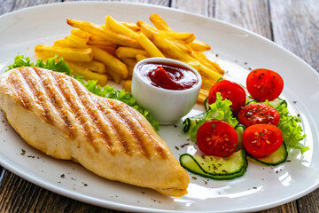 Grilled chicken breast with fries and fresh vegetables on wooden table
