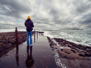 Teenager girl in yellow hat, dark jacket and blue jeans on a foot path to the ocean, dramatic...
