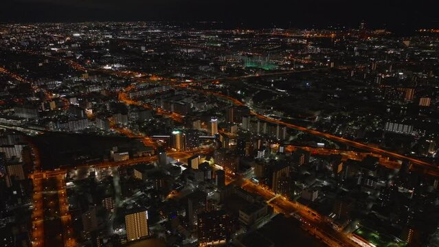 Fly above large city at night. Aerial panoramic view of metropolis with orange illuminated streets and buildings in urban boroughs. Osaka, Japan