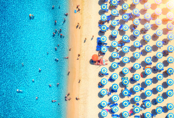 Aerial view of colorful umbrellas on white sandy beach, swimming people in blue sea at sunset in...
