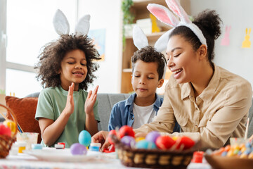Smiling mother and cute kids wearing bunny ears painting easter eggs at home. Happy African...