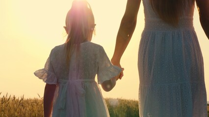 Mother and daughter walking at sunny autumn wheat field holding hands with love tenderness back view closeup. Unrecognizable woman and girl in white dress going together at sunset sunrise fall meadow